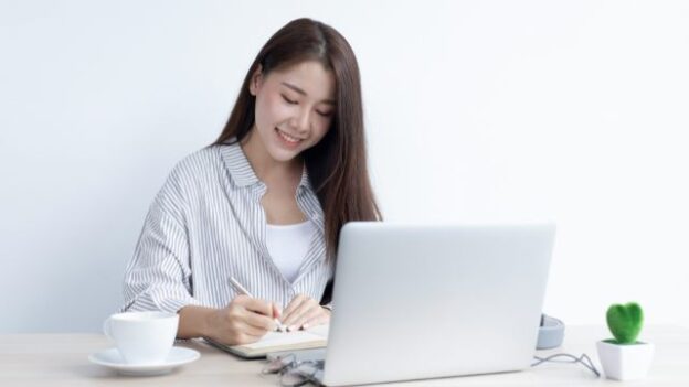 Phụ nữ mỉm cười viết sổ tay trên bàn làm việc với laptop và tách cà phê, không gian sáng sủa và đơn giản.
